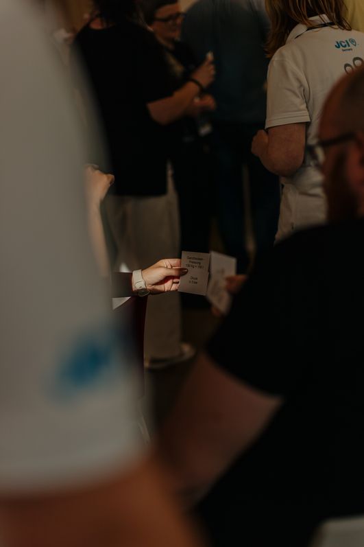 Hand hält Karte bei einer Veranstaltung, unscharfe Personen im Hintergrund