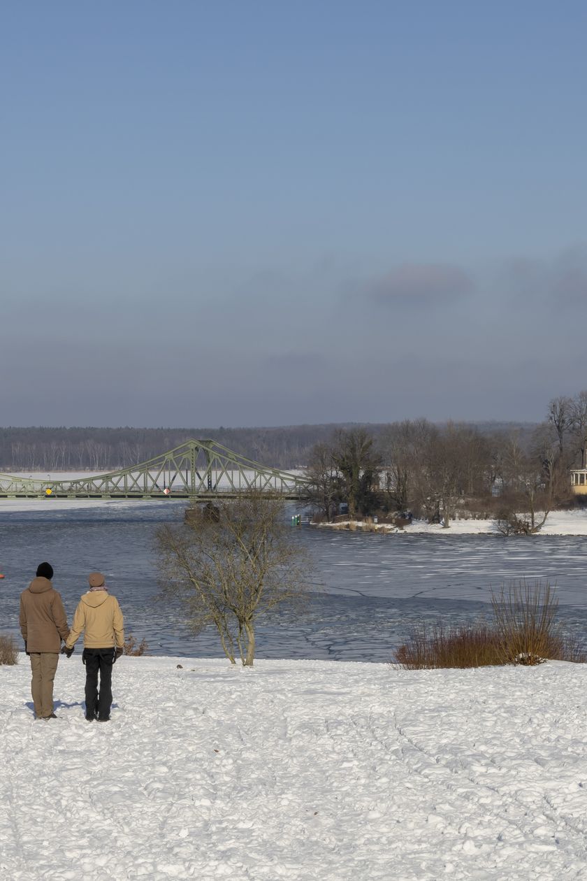 Two people are standing in the wintry Babelsberg Park and looking at the Glienicke Bridge.