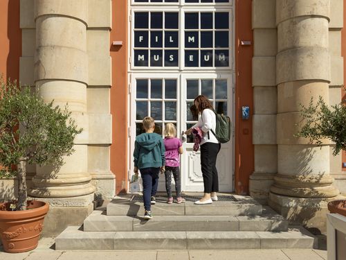 Frau mit zwei Kindern vor dem Eingang des Filmmuseums Potsdam, flankiert von Säulen und Olivenbäumen in Pflanzkübeln.