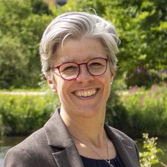 Eine professionell gekleidete Frau steht vor einem Hintergrund aus grünen Blättern und der Nikolaikirche. 