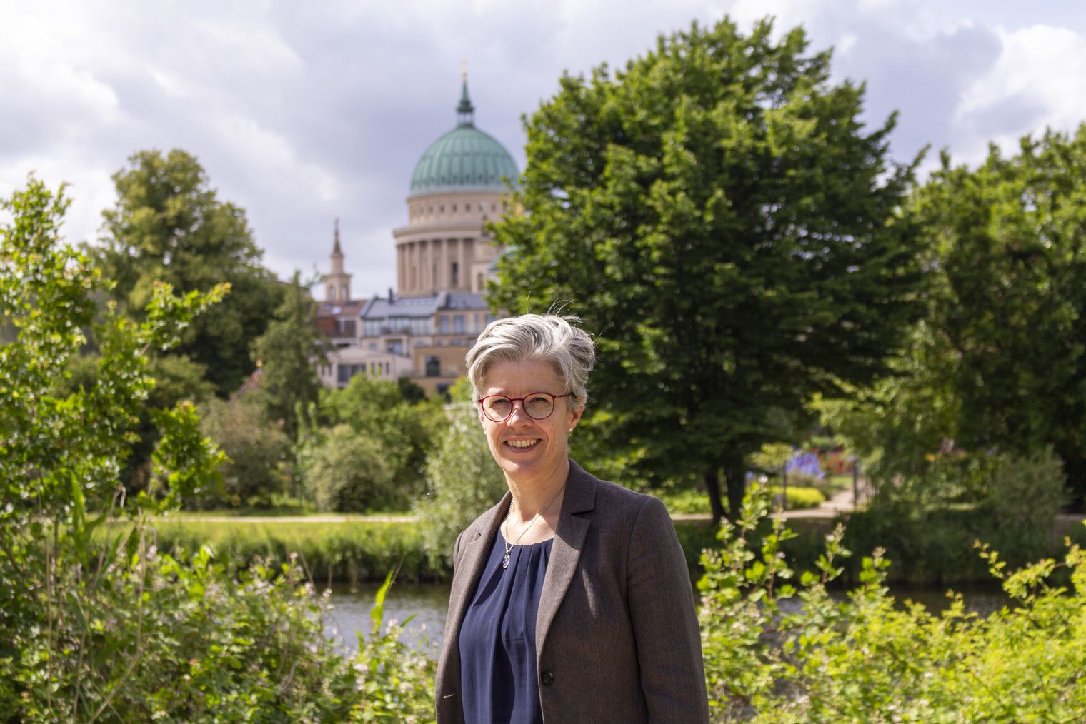 Eine professionell gekleidete Frau steht vor einem Hintergrund aus grünen Blättern und der Nikolaikirche. 