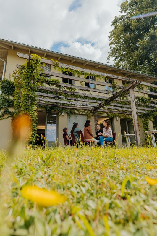 Menschen sitzen auf der Terrasse des Pfingstberghauses unter bewachsener Pergola